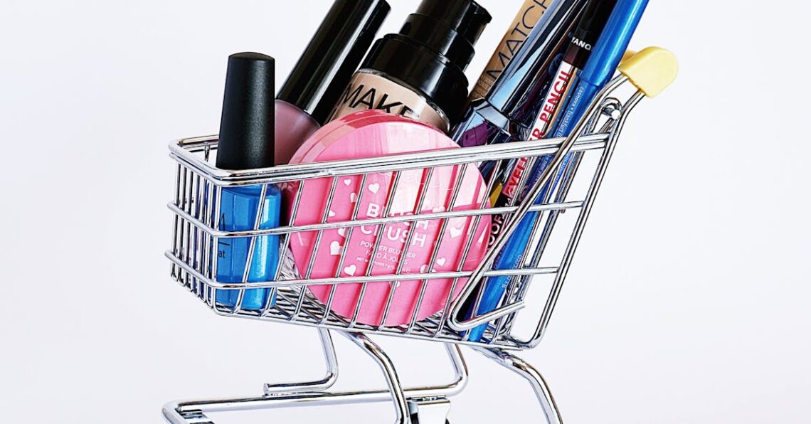 Miniature shopping cart filled with a variety of cosmetics on a white background.